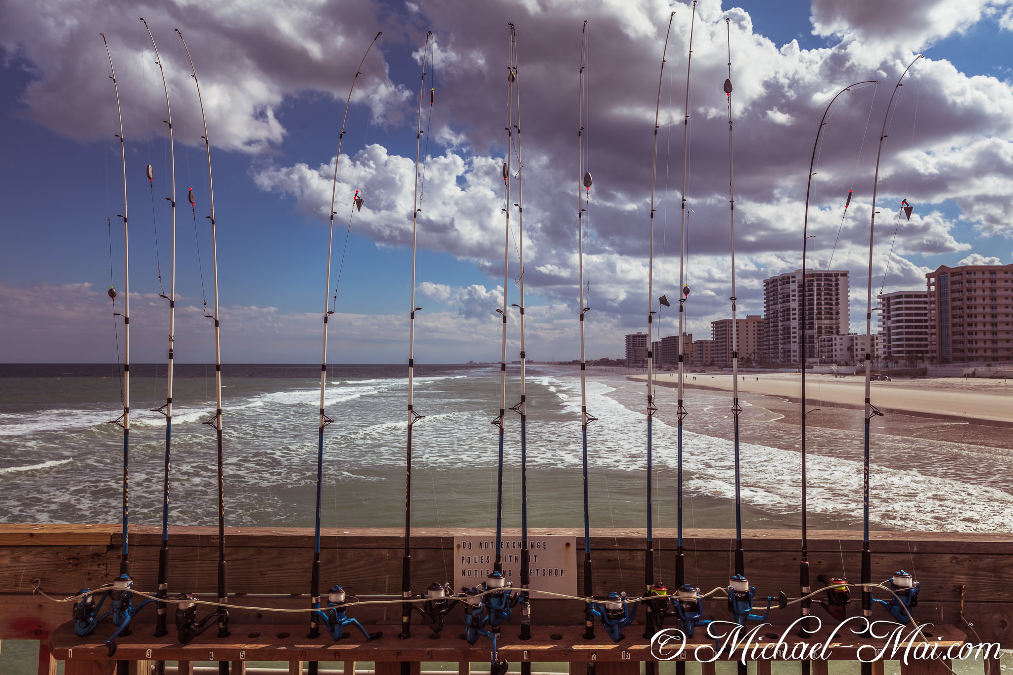Row of fishing rods frames the vibrant ocean and distant beach city. | Daytona Beach Shores, Florida, United States
