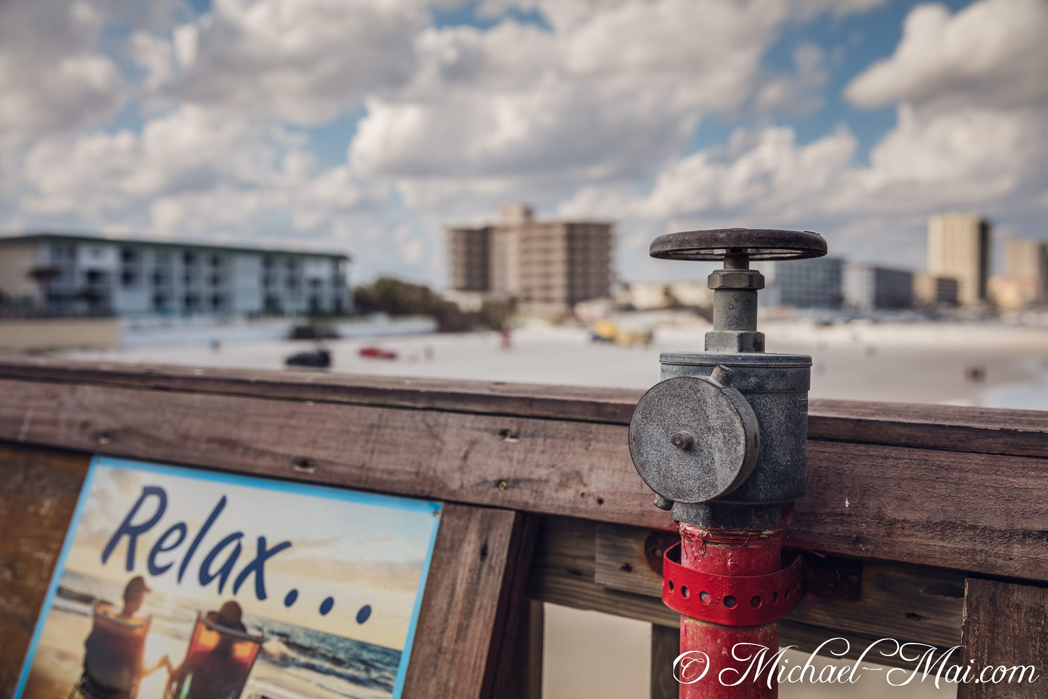 Old valve on rustic railing overlooks the bustling beach, a 'Relax' sign peeking below. | Daytona Beach Shores, Florida, United States