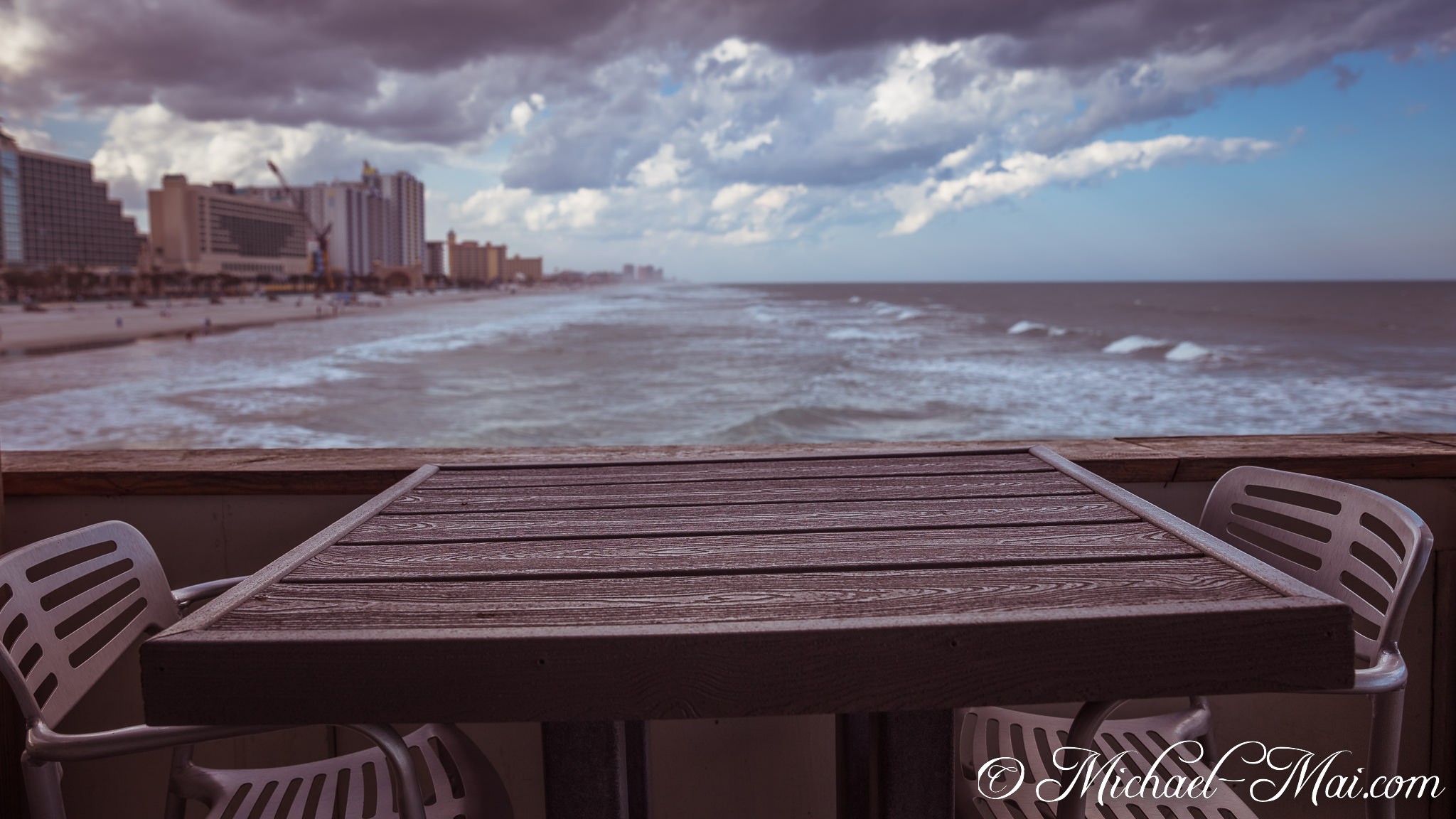 Inviting patio seating overlooks a vibrant, distant coastline under dynamic clouds. | Daytona Beach, Florida, United States