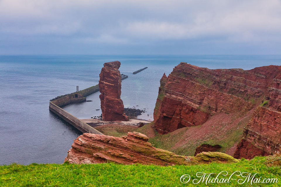 Soaring red rock pillar guards a concrete pier against the expansive, still ocean.