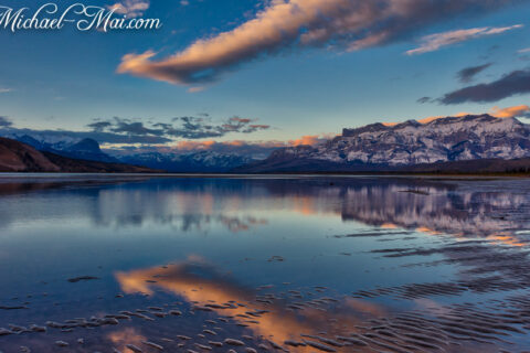 Mirroring mountains reflect a painted sky over rippled shallows at dusk.