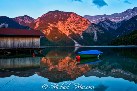 Sun-kissed peaks and a lakeside boathouse reflect on the still alpine lake.