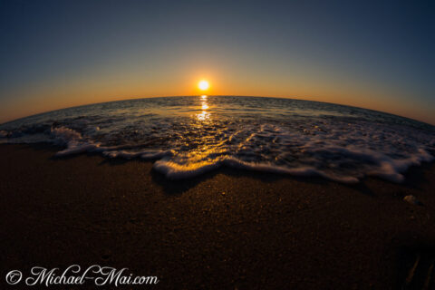 Golden light streaks across the curving ocean, washing over frothy waves at dusk.