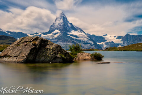 Majestic Matterhorn peaks over a still alpine lake, framed by dramatic clouds and ancient rocks.