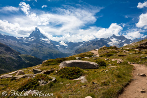Rugged path traverses sun-drenched alpine slopes with the formidable Matterhorn piercing the clouds.