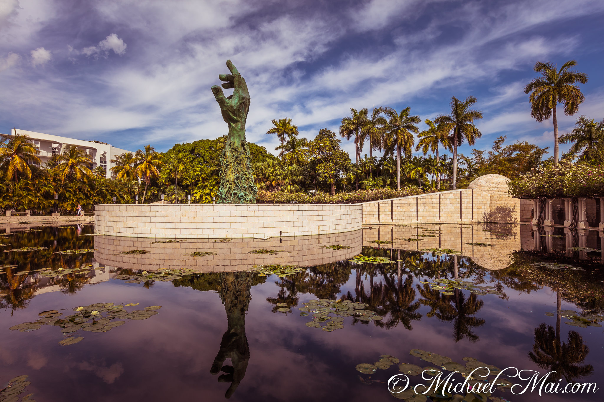 Towering bronze hand dramatically emerges from dark reflective waters under a vivid sky. | Miami Beach, Florida, United States