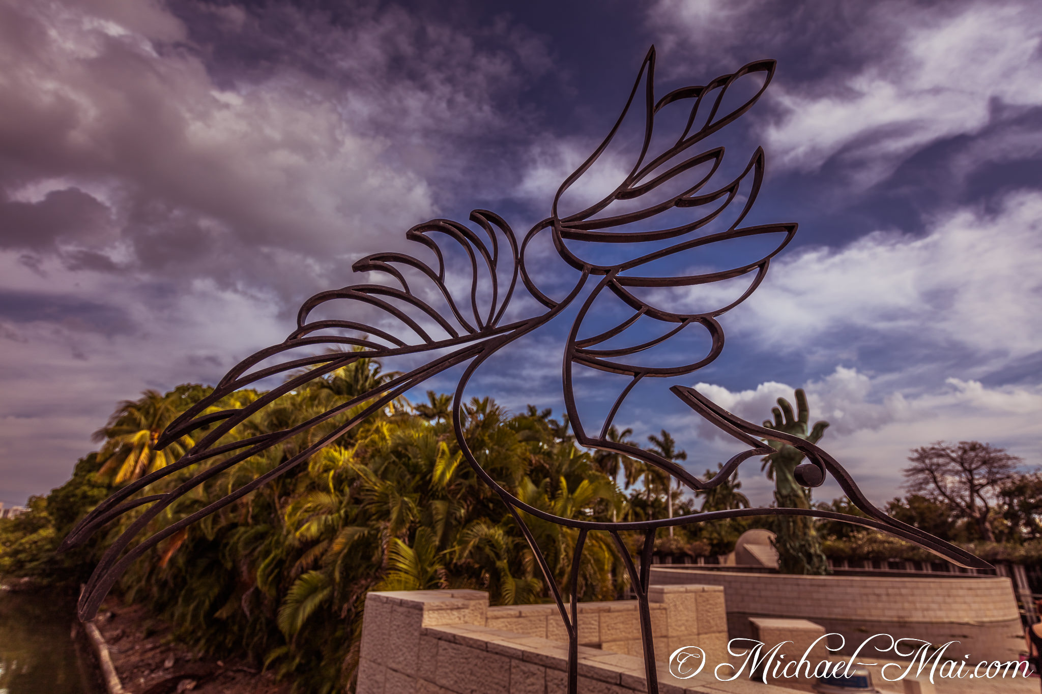 Sleek metal bird sculpture soars, framed by dramatic clouds and vibrant palm trees. | Miami Beach, Florida, United States