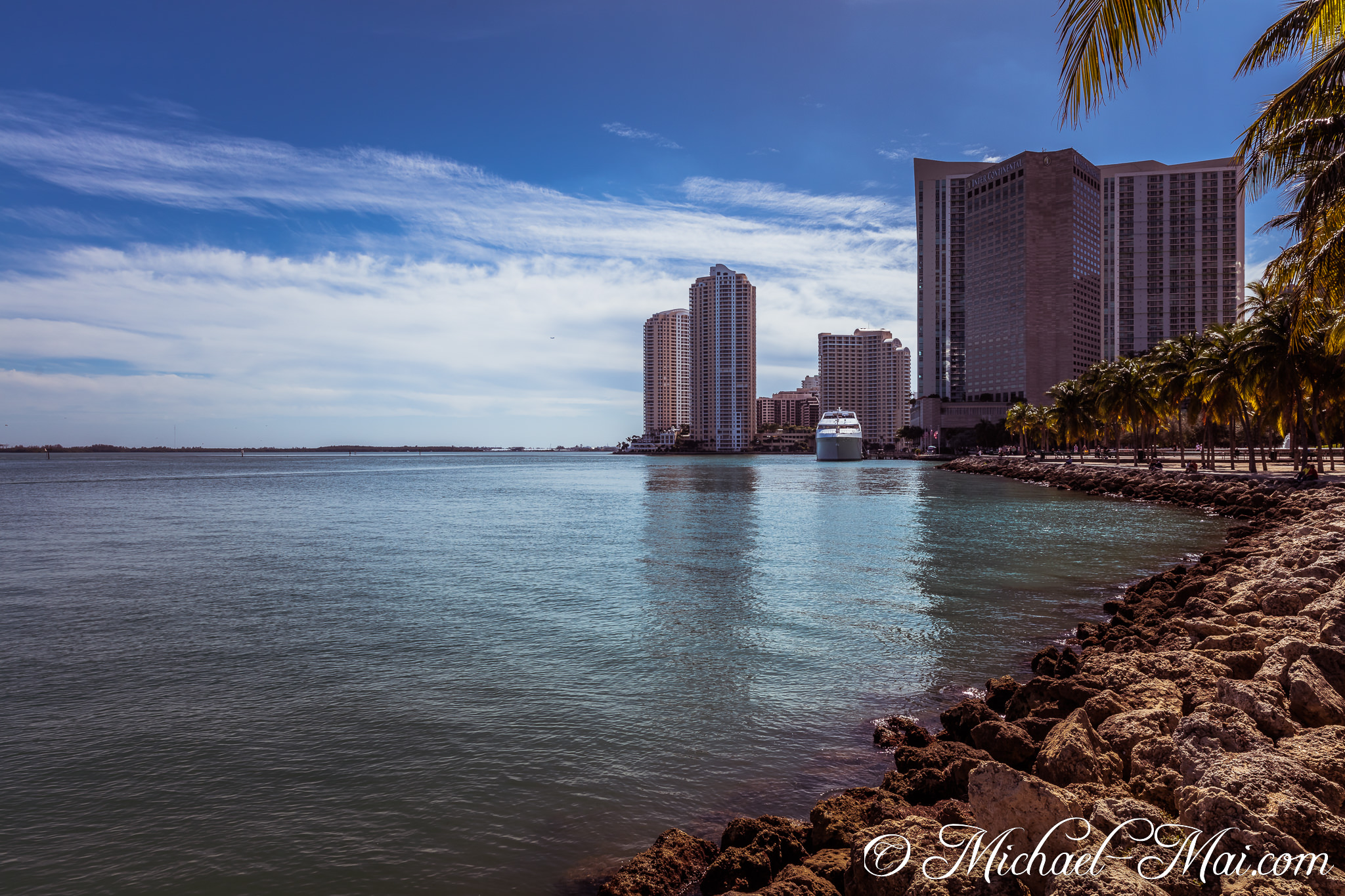 Towering buildings overlook calm bay waters, a vibrant cityscape meeting a rocky tropical shore. | Miami, Florida, United States