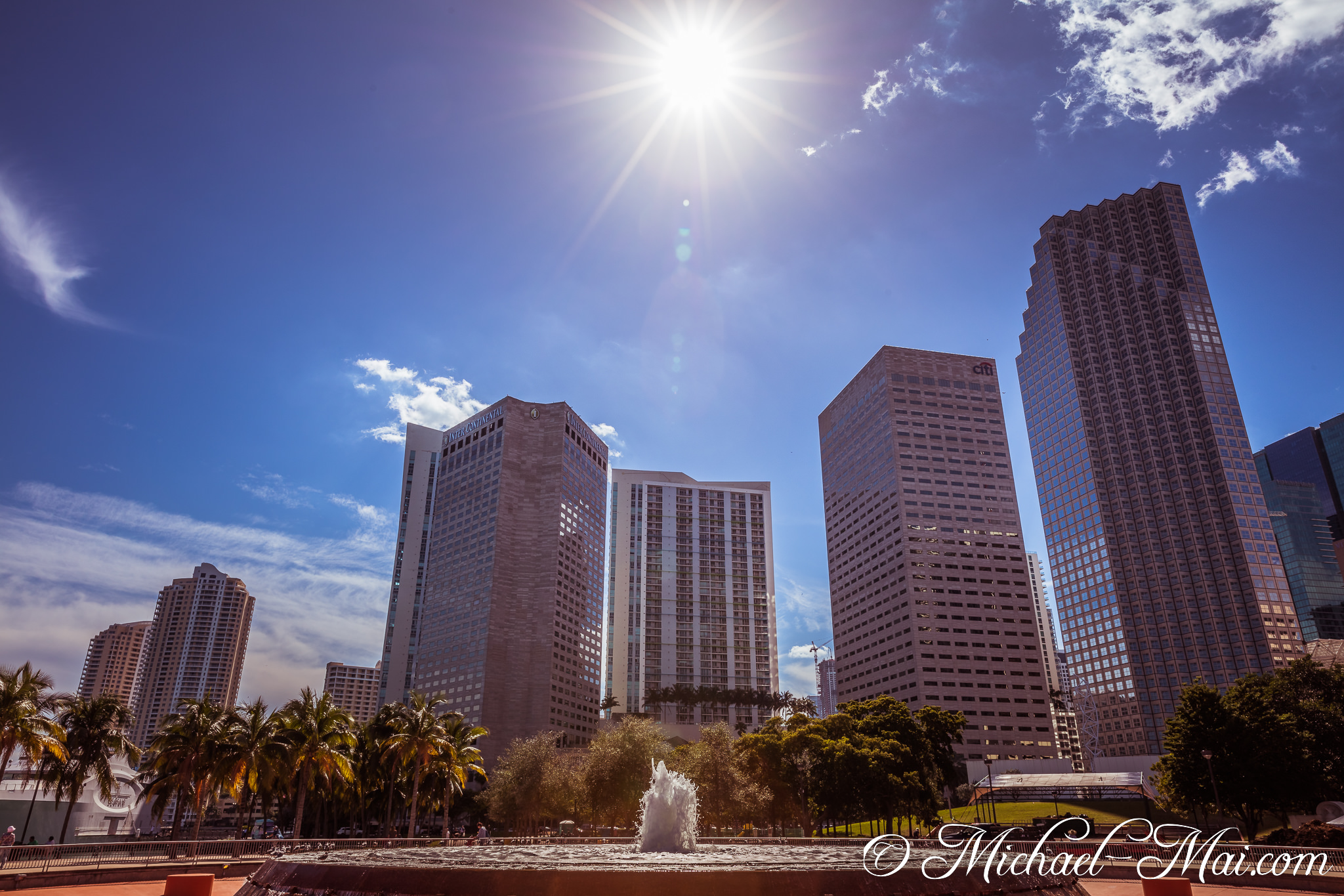 Blinding sun illuminates modern skyscrapers and a lively city fountain. | Miami, Florida, United States