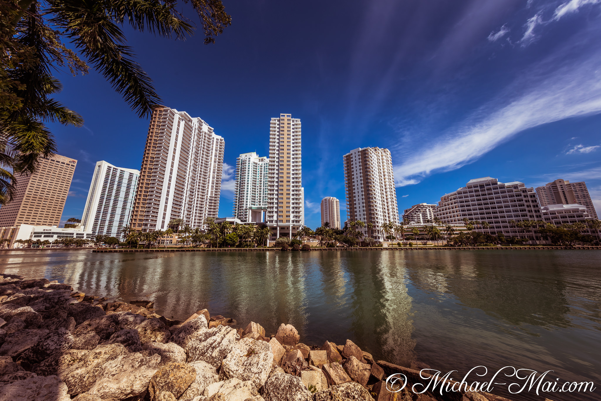 Towering Miami skyscrapers mirrored in the deep bay, a scene anchored by textured rocks. | Miami, Florida, United States