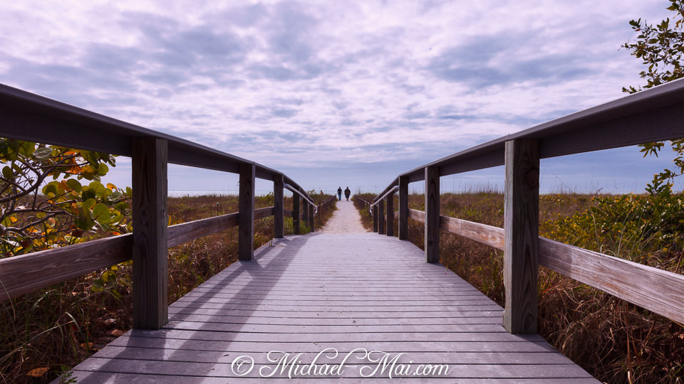 Framed by dune flora, a boardwalk extends to two figures on the beach path.
