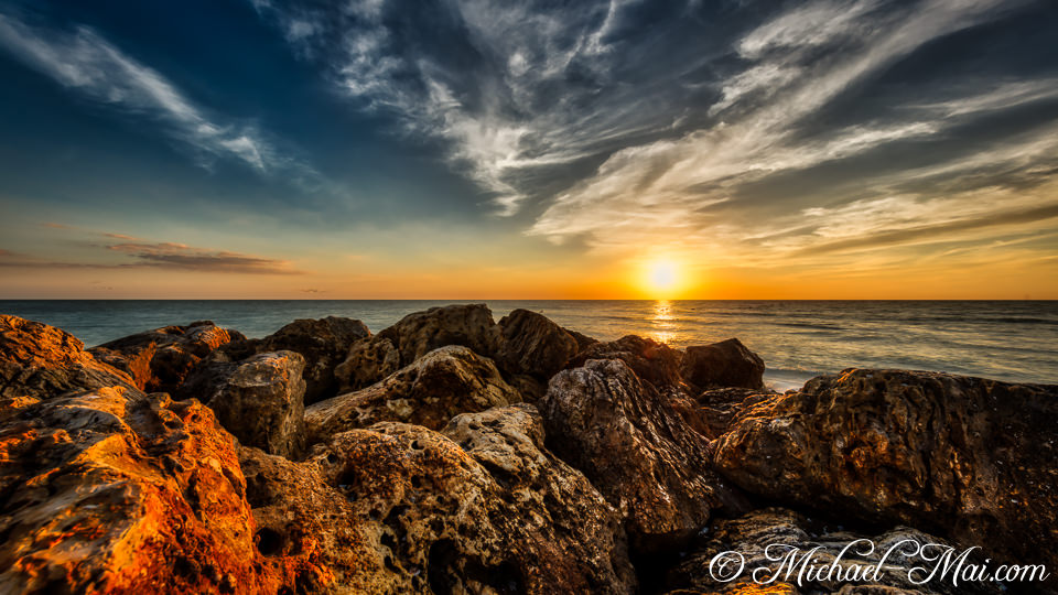 Golden hour light bathes jagged coastal rocks, illuminating a fiery ocean sunset.