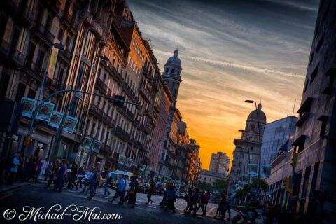 Golden light bathes historic city buildings as pedestrians stream across a vibrant street.