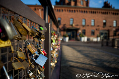 Gleaming and worn locks densely cover a metal fence before an old brick structure.