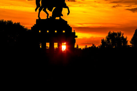The sun bursts through a monument's base, backlighting a grand equestrian statue.