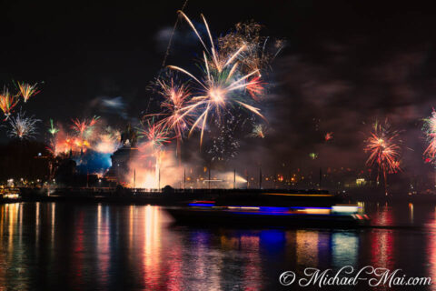 Night explodes with colorful fireworks above the river, a passing boat blurring lights.