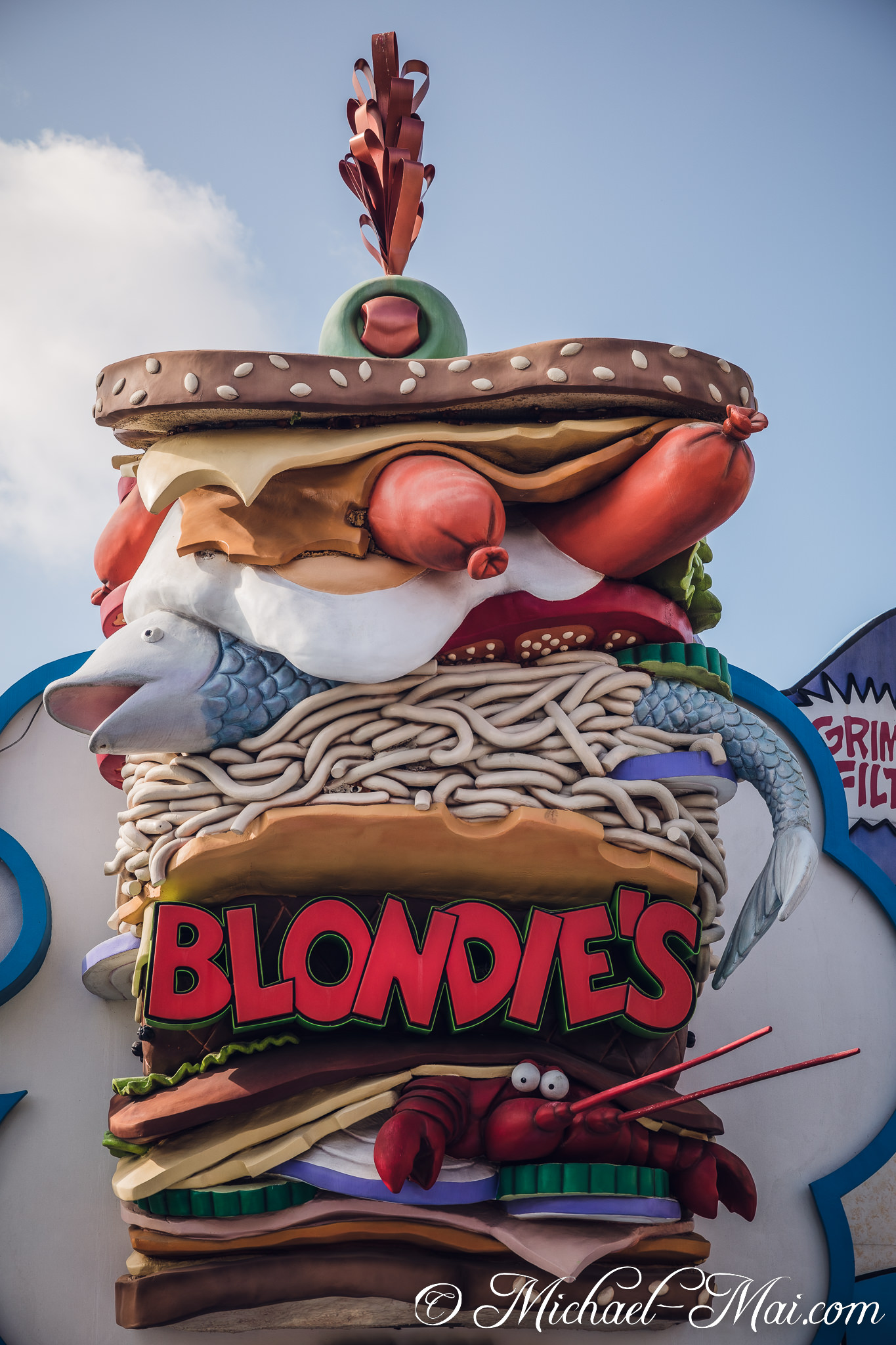 Eclectic ingredients spill from Blondie's gigantic, whimsical sandwich advertisement under blue sky. | Orlando, Florida, United States